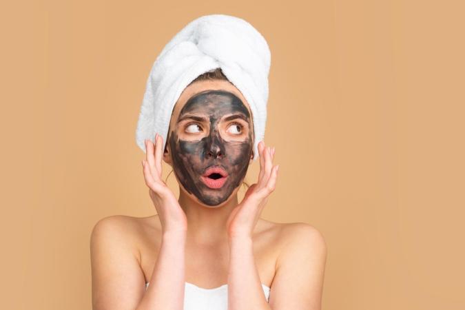 Een vrouw met een gezichtsmasker kijkt verbaasd, met grote ogen en een open mond. Ze houdt haar handen vlak bij haar gezicht, alsof ze geschrokken of verrast is.