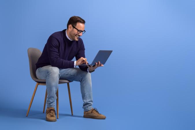 Man zit op een stoel, kijkt lachend naar zijn laptop en typt erop. Zijn gezichtsuitdrukking is vrolijk en geconcentreerd.