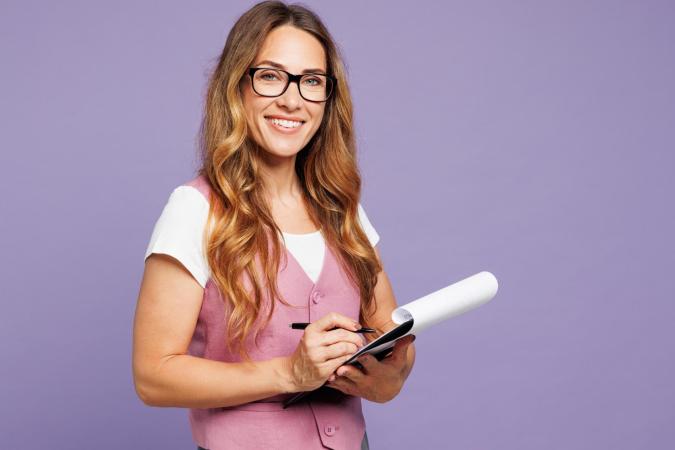 Een vrouw met een vriendelijke glimlach kijkt naar de camera terwijl ze een notitieblok vasthoudt en met een pen iets opschrijft. Ze draagt een bril en heeft lang golvend haar.