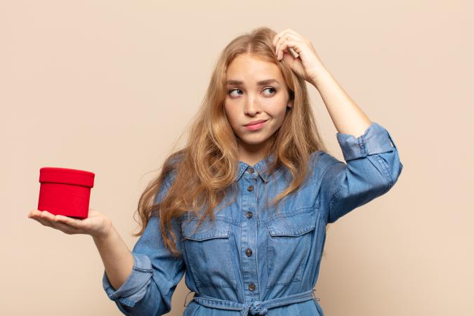 A young woman with long hair wearing a denim dress looks puzzled, scratching her head with one hand while holding a round box in the other hand, as if unsure what to do with it.