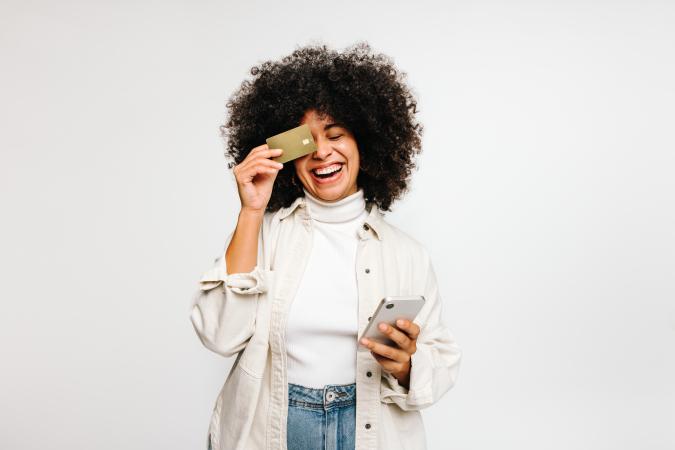 A young woman with curly hair smiles and laughs while holding a credit card up to one eye and a phone in her other hand.