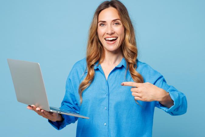 A cheerful woman with long wavy hair holds a laptop in one hand and points to it with the other, smiling confidently 