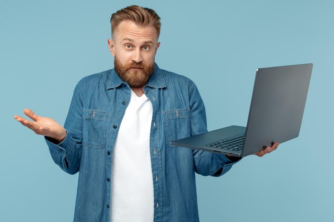A confused man with a beard holds a laptop in one hand while shrugging with the other, expressing uncertainty or puzzlement.