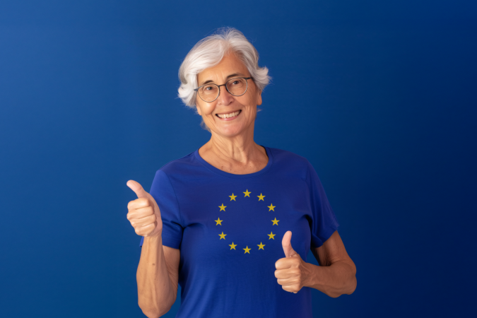 An elderly woman smiling and giving thumbs up, wearing glasses and a t-shirt featuring the European Union flag 