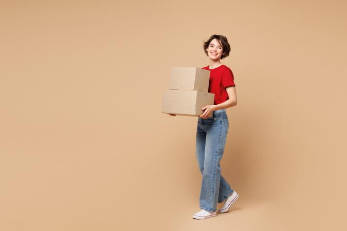 A woman smiling while carrying two stacked cardboard boxes, standing and looking forward.