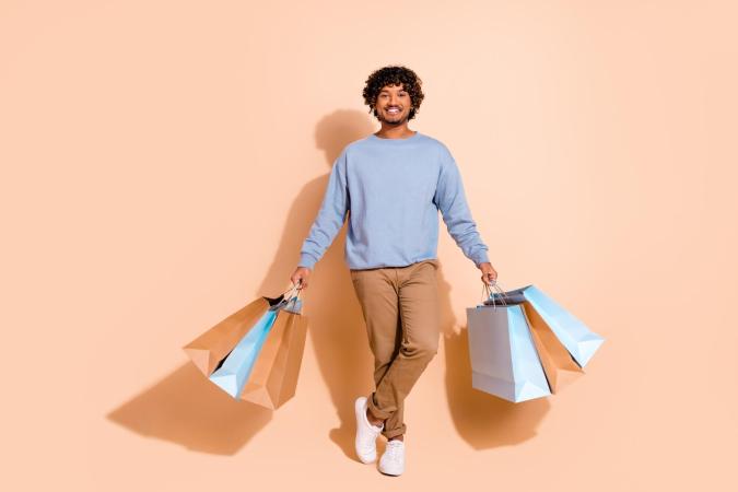 Jonge man met een lichte glimlach, staat ontspannen met één been gekruist en houdt in elke hand meerdere boodschappentassen vast. Zijn houding straalt tevredenheid en voldoening uit.