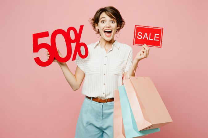 A cheerful young woman holding several shopping bags in one hand and red sale signs showing "50%" and "SALE" in the other, expressing excitement and enthusiasm.