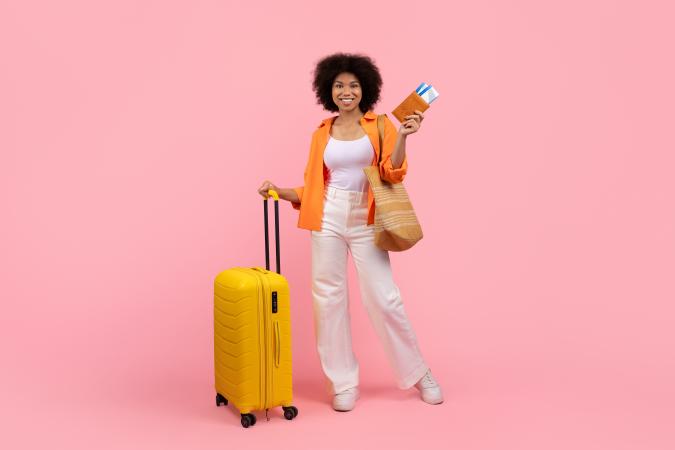 A woman stands smiling confidently while holding a suitcase with one hand and a wallet with travel tickets in the other hand, ready for a trip.