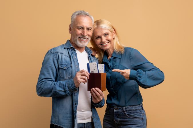 A smiling older man is holding a wallet with boarding passes inside, while a woman next to him is pointing at the wallet with an excited expression.