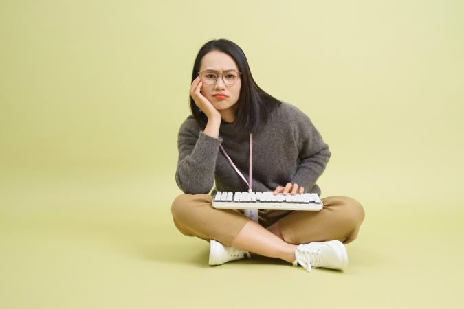 Vrouw zit met gekruiste benen op de grond, kijkt met een frons en licht beteuterde mond naar de camera. Ze ondersteunt haar hoofd met één hand en de andere hand rust op een toetsenbord dat op haar schoot ligt.