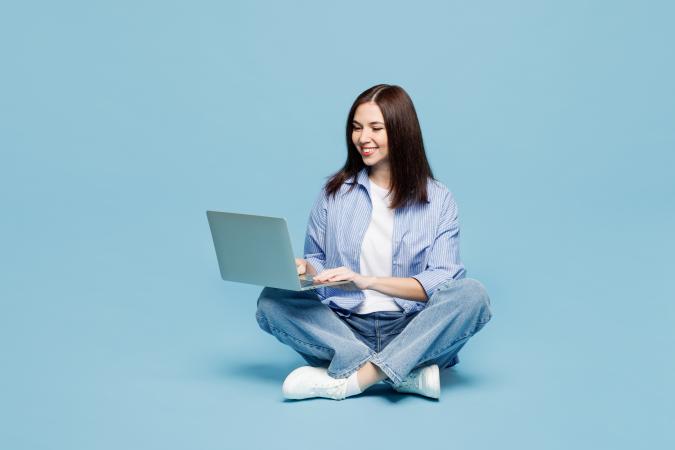 A woman sitting cross-legged on the floor, smiling while working on a laptop placed on her lap.