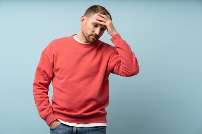 A man wearing a sweater looks worried or stressed as he holds one hand on his forehead and the other in his pocket.
