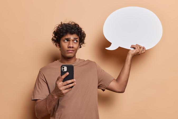 A man looks sideways with a doubtful or skeptical expression, holding a smartphone in one hand and a blank speech bubble sign in the other.