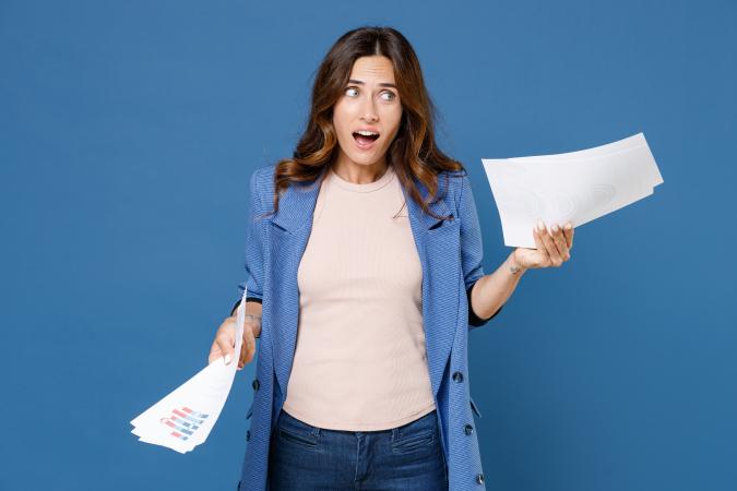 Vrouw kijkt verbaasd en bezorgd terwijl ze papieren in beide handen vasthoudt, met haar mond open en haar blik naar links gericht.