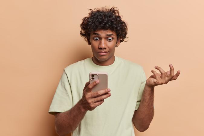 A young man looking confused and frustrated, holding a smartphone in one hand and raising the other hand with fingers spread, as if questioning something on the phone.