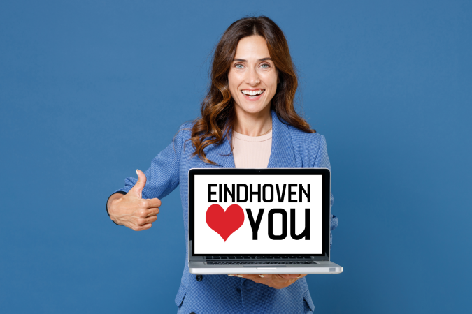 A smiling woman in a blazer holds a laptop showing the message EINDHOVEN LOVES YOU and gives a thumbs-up