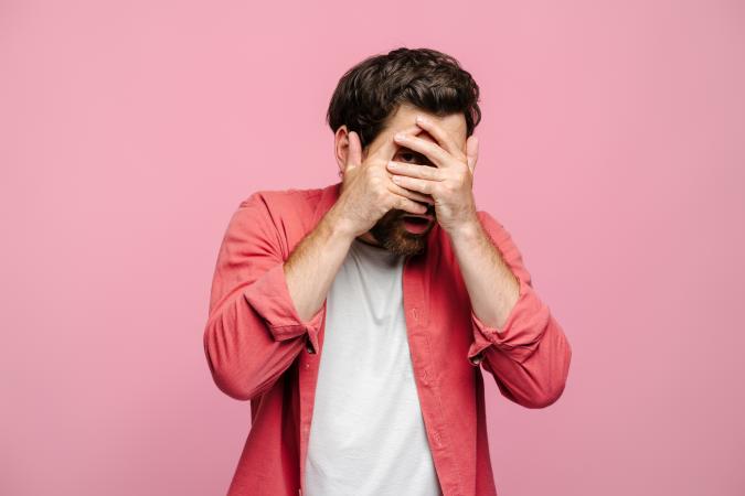 Man kijkt angstig en kijkt door zijn vingers. Hij draagt een rood shirt en staat voor een effen roze achtergrond.