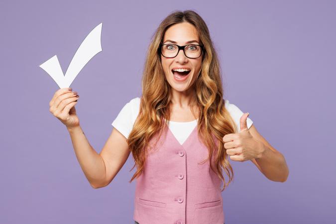 A woman with long hair and glasses smiles widely, holding a large paper checkmark in one hand and giving a thumbs-up with the other, expressing approval and positivity.