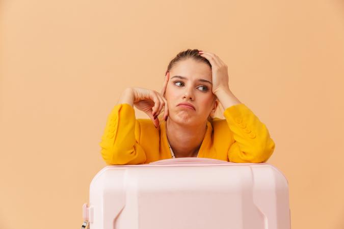Woman with orange blazer leaning on pink suitcase, waiting for delayed flight. Peach-coloured background