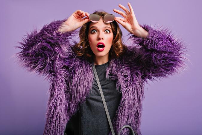 Vrouw met rode lipstick en paars jasje op paarse achtergrond kijkt verbaasd, houdt zonnebril omhoog