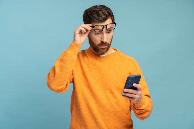 Man in orange jumper looks at phone and raises glasses when reading bankruptcy announcement - solid light blue background