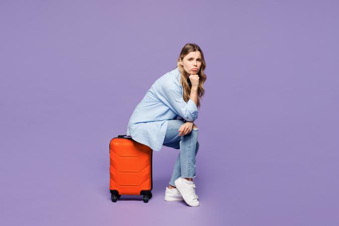 Vrouw in jeans en lichtblauwe blouse zit op rode koffer en wacht op de volgende vlucht (effen paarse achtergrond)